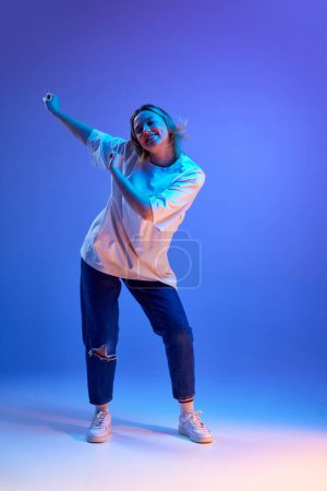 Photo for Full-length portrait of beautiful young girl in casual white t-shirt cheerfully dancing against blue studio background in neon light. Concept of youth, human emotions, casual fashion - Royalty Free Image