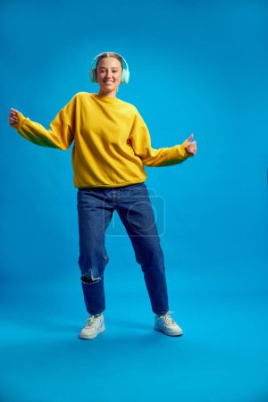 Photo for Full-length image of young girl in yellow sweatshirt listening to music in headphones and dancing against blue studio background. Concept of youth, human emotions, casual fashion - Royalty Free Image
