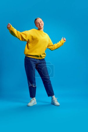 Photo for Full-length image of young girl in yellow sweatshirt and casual jeans dancing, having fun against blue studio background. Concept of youth, human emotions, casual fashion - Royalty Free Image