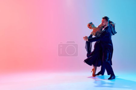 Foto de Hermoso hombre joven y mujer guapa, bailarines de salón haciendo actuación, bailando contra el degradado fondo azul rosado en luz de neón. Concepto de clase de baile, hobby, arte, escuela de baile, talento - Imagen libre de derechos