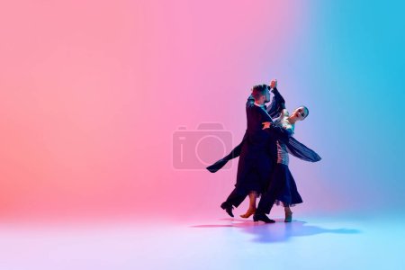 Foto de Joven hombre y mujer, talentosos bailarines de salón en movimiento, bailando con trajes negros contra el degradado fondo azul rosado en luz de neón. Concepto de clase de baile, hobby, arte, escuela de baile, talento - Imagen libre de derechos