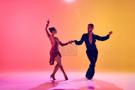Foto de Bailarinas masculinas y femeninas realizando una danza latina con ropa elegante contra el degradado fondo amarillo rosado en luz de neón. Concepto de clase de baile, hobby, arte, escuela de baile, talento - Imagen libre de derechos