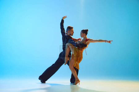 Foto de Joven hombre y mujer en elegantes trajes escénicos realizando samba, bailando sobre fondo azul degradado en luz de neón. Concepto de clase de baile, hobby, arte, escuela de baile, talento - Imagen libre de derechos