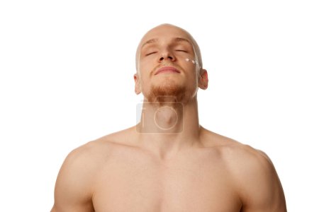 Photo for Shirtless, muscular young bald man with beard standing with eyes closed and moisturizing cream of face isolated against white studio background. Concept of skin care, male beauty and cosmetics, youth - Royalty Free Image