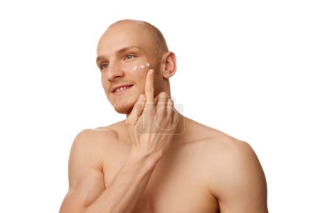 Photo for Handsome bald shirtless young man with muscular body, applying moisturizing face cream isolated against white studio background. Concept of skin care, male beauty and cosmetics, youth, spa, self-care - Royalty Free Image