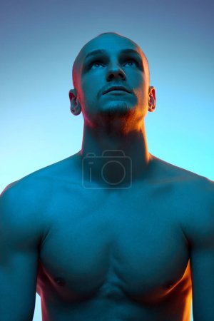 Photo for Portrait of young bald man in his 20s with muscular shirtless body sitting and looking upwards against blue background in neon light. Concept of male beauty, sportive and healthy lifestyle, body care - Royalty Free Image