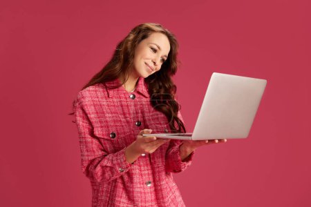 Téléchargez les photos : Jeune belle femme tenant un ordinateur portable, travaillant en ligne, faisant des projets, communiquant via Internet sur fond de studio rose. Concept de jeunesse, mode de vie, mode décontractée, émotions humaines - en image libre de droit