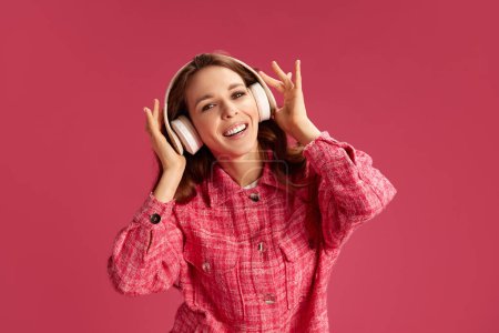 Foto de Mujer joven feliz, hermosa y sonriente con camisa a cuadros, sonriendo. escuchar música en auriculares sobre fondo de estudio rosa. Concepto de juventud, estilo de vida, moda casual, emociones humanas - Imagen libre de derechos
