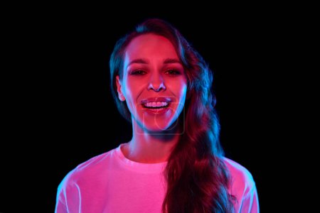 Photo for Portrait of beautiful, happy young woman with nude makeup, curly hair posing, smiling against black studio background in neon light. Concept of youth, lifestyle, casual fashion, human emotions - Royalty Free Image