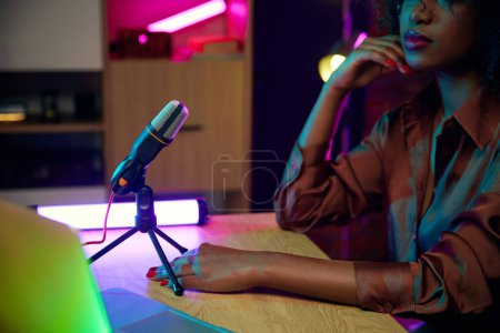 Téléchargez les photos : Concentre-toi sur le micro. Image recadrée d'une jeune femme africaine assise à table avec un appareil d'enregistrement, un microphone et un ordinateur portable et faisant du podcast. Communication en ligne, technologie moderne, médias de masse - en image libre de droit
