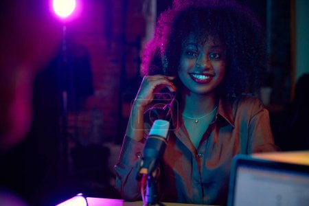 Foto de Joven mujer africana sonriente, influyente de las redes sociales, bloguera sentada con micrófono en una entrevista de podcast. Haciendo show. Concepto de comunicación en línea, tecnología moderna, medios de comunicación - Imagen libre de derechos