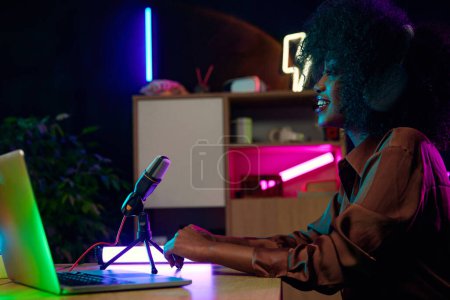 Téléchargez les photos : Jeune femme africaine souriante assise au bureau à la maison dans un casque, avec microphone et ordinateur portable, enregistrement podcast en ligne. Entreprises de médias sociaux. Concept de journalisme, technologie moderne, médias de masse - en image libre de droit