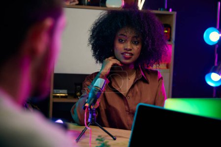 Téléchargez les photos : Jeune femme africaine, journaliste, blogueur enregistrement podcast dans le bureau à domicile, faire une interview, discuter de sujets de style de vie intéressants. Concept de communication en ligne, technologie moderne, médias de masse - en image libre de droit