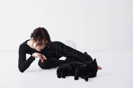 Téléchargez les photos : Lien entre humain et animal de compagnie. Belle jeune femme en vêtements noirs jouant avec chat noir sur fond de studio blanc. Concept de l'être humain et des animaux, connexion, espace acceptant les animaux - en image libre de droit
