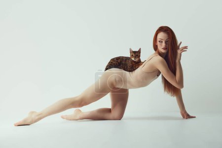 Photo for Femininity and self-identity. Passionate, beautiful young redhead woman with slim body with purebred Bengal cat lying on back over white studio background. Concept of animal theme, bonding, wellness - Royalty Free Image