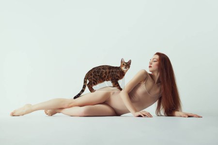 Photo for Beautiful, young, elegant redhead woman in beige bodysuit lying with purebred Bengal at against white studio background. Concept of pet companion, animal care, well-being, beauty and fashion - Royalty Free Image