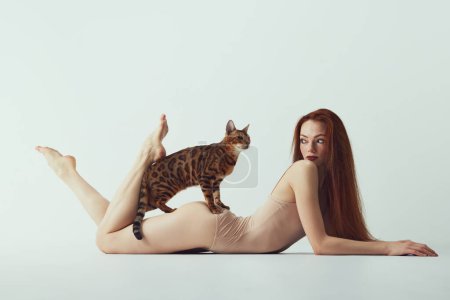 Photo for Passion and tenderness. Young redhead beautiful girl with long hair, slim body lying on floor with purebred Bengal cat against white studio background. Concept of beauty, animal theme - Royalty Free Image