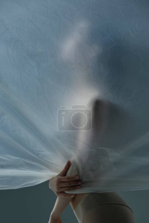 Téléchargez les photos : Silhouette de jeune femme posant nue avec un matériau transparent, mat et texturé sur fond gris. Concept d'esthétique corporelle, féminité, beauté, santé, art - en image libre de droit