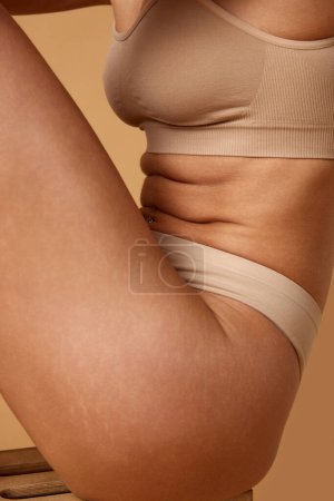 Téléchargez les photos : Vue latérale rapprochée de l'abdomen des femmes en sous-vêtements beige, mettant en valeur la texture de la peau et s'adaptant sur fond studio couleur sable. Concept de beauté, procédures de spa, traitements dermatologiques. - en image libre de droit