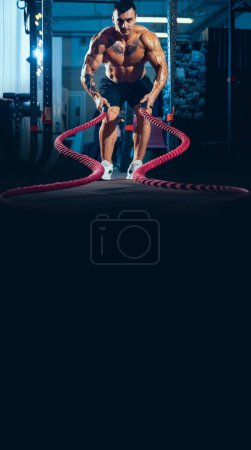 Photo for Young healthy man, athlete doing exercise with the ropes in the gym. Male model training hard his upper body. Vertical image. Concept of fitness, sport, workout, athlete, power and health - Royalty Free Image