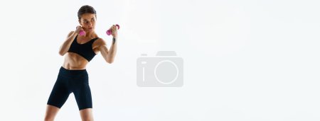 Photo for Young woman with sportive body training, doing uppercut with dumbbells isolated over white studio background. Concept of sport, health and body care, fitness app, exercises templates. Banner - Royalty Free Image