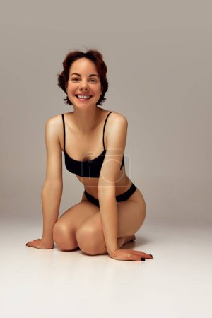 Photo for Portrait of happy smiling young woman with slim body posing in black underwear against beige studio background. Concept of female health care, sexuality, passion and pleasure, wellness - Royalty Free Image
