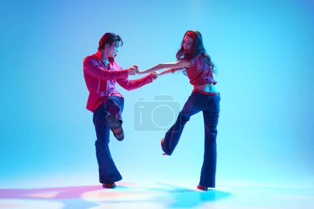 Pareja joven positiva, activa, hombre y mujer en ropa elegante bailando danza retro sobre fondo azul en luz de neón. Concepto de pasatiempo, clase de baile, fiesta, 50, cultura de los 60, juventud