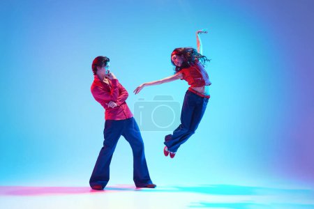 Hermosa joven bailando en pose dinámica mientras el hombre mira con admiración sobre fondo azul en luz de neón. Baile retro. Concepto de pasatiempo, clase de baile, fiesta, 50, cultura de los 60, juventud