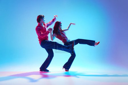 Foto de Feliz, positivo joven hombre y mujer en ropa retro bailando boogie woogie baile retro sobre fondo azul en luz de neón. Concepto de pasatiempo, clase de baile, fiesta, 50, cultura de los 60, juventud - Imagen libre de derechos