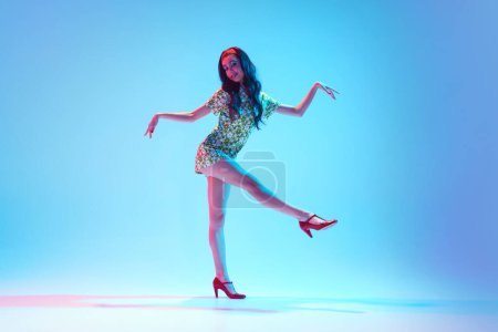 Foto de Chica joven artística, talentosa y hermosa en vestido elegante bailando sobre fondo azul en luz de neón. Concepto de pasatiempo, clase de baile, fiesta, 50, cultura de los 60, juventud - Imagen libre de derechos
