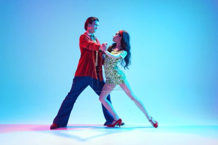Photo for Artistic, talented, positive young man and woman, stylish couple dancing retro dance against blue background in neon light. Concept of hobby, dance class, party, 50s, 60s culture, youth - Royalty Free Image