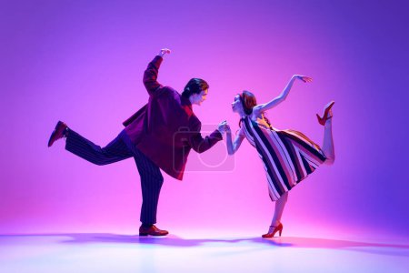 Téléchargez les photos : Élégant, beau jeune couple, homme et femme dans des vêtements rétro danse swing sur fond violet dans la lumière au néon. Concept de passe-temps, cours de danse, fête, années 50, 60 culture, jeunesse - en image libre de droit