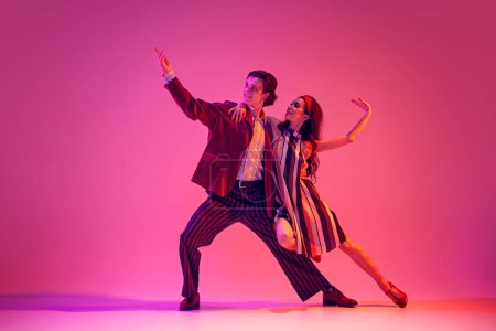 Künstlerischer junger Mann und elegante Frau, Paar tanzen Retro-Tanz in stilvollen Kostümen vor rosa Hintergrund in Neonlicht. Konzept von Hobby, Tanzkurs, Party, Kultur der 50er, 60er Jahre, Jugend