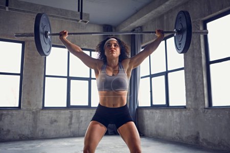 Front view of focused young athlete woman in sportswear doing squat technique with barbell overhead in motion at gym. Concept of professional sport, active lifestyle, bodybuilding, action. Ad