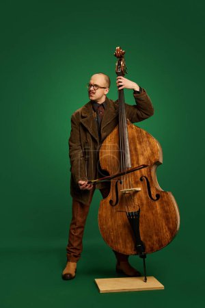 Photo for Man with stylish moustache in brown suit playing double bass against green studio background. Classical music event. Concept of music, performance, art, entertainment, festival, performance, ad - Royalty Free Image