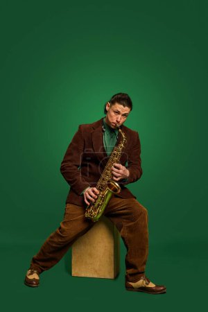 Téléchargez les photos : Musicien masculin en costume classique marron jouant du saxophone sur fond de studio vert foncé. Concert de jazz. Concept de musique, performance, art, divertissement, festival, performance, annonce - en image libre de droit