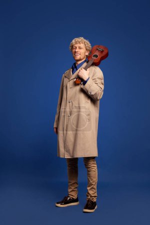 Photo for Smiling man in stylish trench coat standing with ukulele against dark blue background. Creative solo performance. Concept of music, performance, art, entertainment, festival, performance, ad - Royalty Free Image