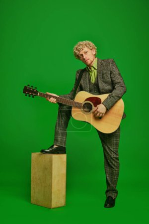 Curly young man in a suit playing guitar against green studio background. Poster for live music event. Concept of music, performance, art, entertainment, festival, performance, ad