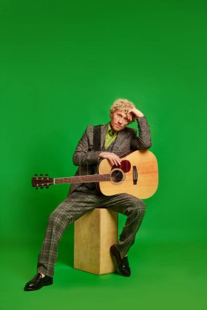 Photo for Passionate musician. Curly young man in a suit sitting with guitar against green studio background. Live music event. Concept of music, performance, art, entertainment, festival, performance, ad - Royalty Free Image