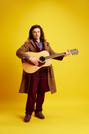 Photo for Man with long hair, musician in coat playing guitar against bright yellow background. Performer. Concept of music, performance, art, entertainment, festival, performance, ad - Royalty Free Image