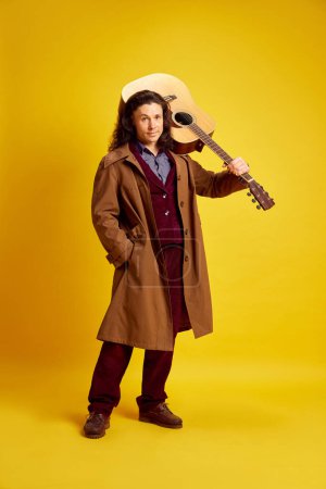 Man with long hair, musician in coat standing with guitar against bright yellow background. Solo performer. Concept of music, performance, art, entertainment, festival, performance, ad