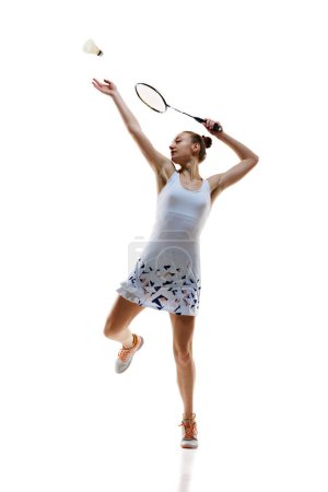 Full-length image of motivated athlete in motion, young girl, badminton playing hitting shuttlecock in jump isolated over white background. Concept of professional sport, active lifestyle, hobby, game