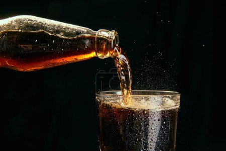 Téléchargez les photos : Soda, coke, bière noire coulant de la bouteille dans du verre sur fond noir. Concept de boissons alcoolisées et non alcoolisées, rafraîchissements. Affiche, bannière pour la publicité - en image libre de droit