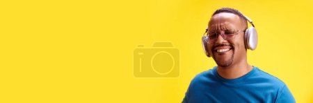 Photo for Smiling African-American man listening to music in headphones against yellow studio background. Concept of human emotions, casual fashion, lifestyle. Banner. Empty space for text, ad - Royalty Free Image