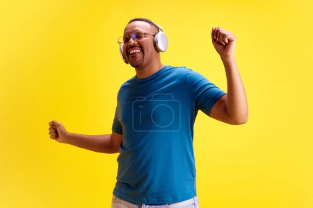 Photo for Cheerful, positive, relaxed African-American man in casual clothes listening to music in headphones and dancing against yellow studio background. Concept of human emotions, casual fashion, lifestyle - Royalty Free Image