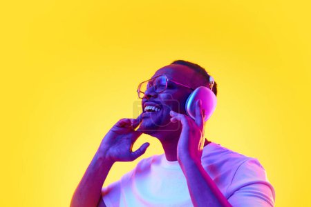 Photo for Portrait of positive African-American man listening to music in headphones and enjoying against yellow studio background in neon light. Concept of human emotions, casual fashion, lifestyle - Royalty Free Image