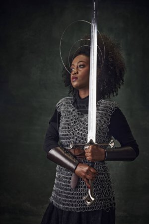 Téléchargez les photos : Élégante jeune femme afro-américaine en armure de maille médiévale avec des anneaux halo au-dessus de la tête tenant l'épée sur fond vert foncé. Concept d'histoire, de beauté et de mode, comparaison des époques - en image libre de droit