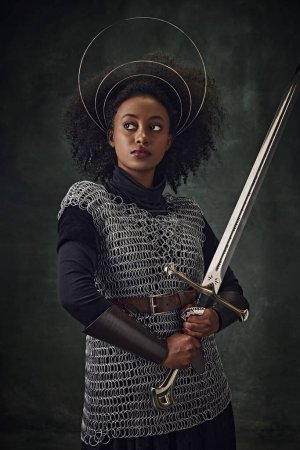 Photo for Serious, young African woman, medieval warrior in chainmail armor with halo-like rings above head and sword against vintage green background. Concept of history, beauty and fashion, comparison of eras - Royalty Free Image