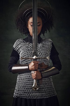 Téléchargez les photos : Portrait de jeune femme africaine, guerrière médiévale en armure de maille avec des anneaux halo au-dessus de la tête tenant l'épée près du visage sur fond vintage foncé. Concept d'histoire, comparaison des époques - en image libre de droit