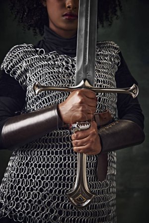 Photo for Close-up of African woman in chainmail holding sword vertically in front of face against vintage green background. Focus on hands and sword. Concept of history, beauty and fashion, comparison of eras - Royalty Free Image